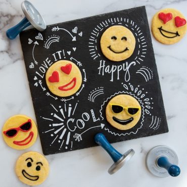 Three baked emoji stamped cookies on chalkboard with illustrations and piped frosting on cookies.