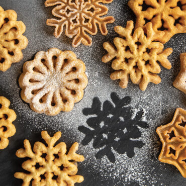 Golden fried rosettes dusted with powdered sugar on dark surface, one piece removed showing circular outline