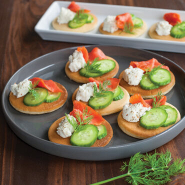 Small golden pancakes on white platter topped with smoked salmon, cucumber slices, dill and cream cheese