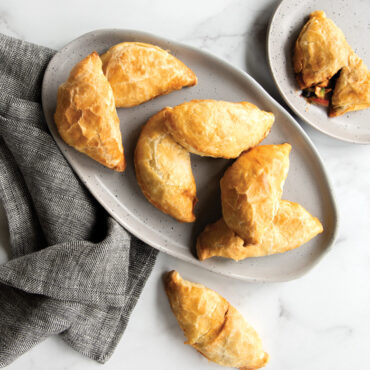 Golden-brown baked empanadas arranged on white platter with gray kitchen towel, showing flaky crimped edges