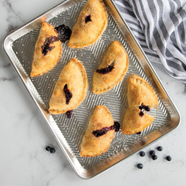 Four golden-brown blueberry pocket pies on Nordic Ware Prism aluminum baking sheet, sprinkled with crystal sugar