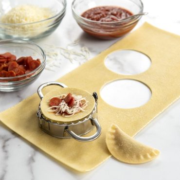 Assembling dumpling maker with pizza fixings, ingredients in bowls in background, product in use.