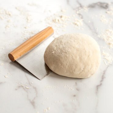 Scraper underneath mound of dough on floured surface.