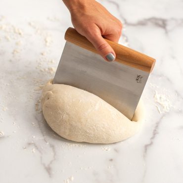 Dough scraper being used to cut  mound of dough in half with end side.