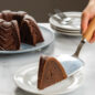 Hand serving slice of chocolate Bundt cake on plate using caker server, cut baked Bundt in background.