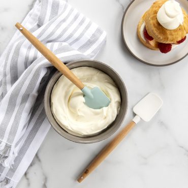 Small spatula in bowl of frosting, white spatula on surface with a plate of biscuit and fruit and whipped frosting on top.