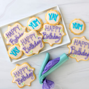 Assorted decorated sugar cookies on white platter with Nordic Ware E-Z Deco icing pen, showing frosted text and designs