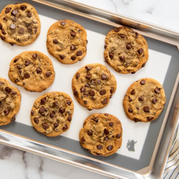 Golden-brown chocolate chip cookies arranged on Nordic Ware red silicone baking mat in aluminum sheet pan