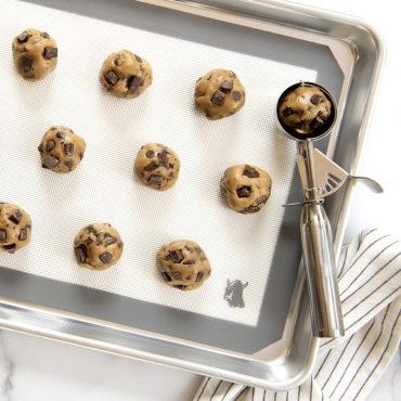 Raw cookie dough scooped onto Deluxe mat with cookie scoop, on baking sheet with towel on surface.