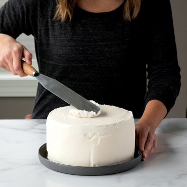 Woman using Straight Icing spatula to frost top of tiered cake, white frosting.