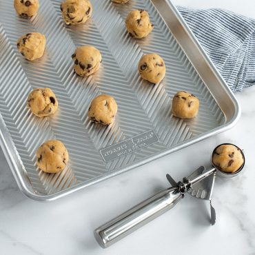 Small cookie scoop filled with chocolate chip cookie dough, pan filled with dough balls.