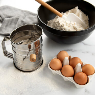 Flour sifter filled with flour next to mixing bowl filled with sifted flour, 6 eggs in egg holder.
