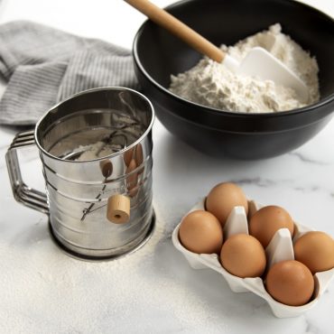 Flour sifter filled with flour next to mixing bowl filled with sifted flour, 6 eggs in egg holder.