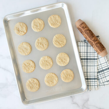 Nordic Ware wooden embossing rolling pin with woodland cottage design next to golden baked cookies on aluminum half sheet pan
