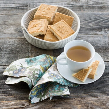 Golden baked shortbread squares with traditional pattern in wicker basket beside white teacup and saucer