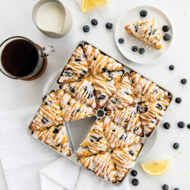 Nordic Ware mini scone pan with six golden blueberry scones drizzled with white glaze, fresh blueberries scattered around