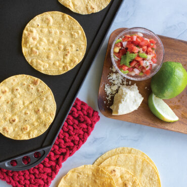 Golden corn tortilla cooking on Nordic Ware aluminum reversible griddle with bowls of salsa and cheese nearby