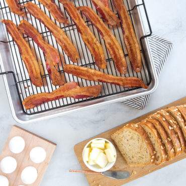Crispy cooked bacon strips on Nordic Ware Extra Large Oven Crisp aluminum baking tray with sliced bread nearby
