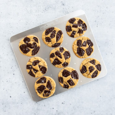 Golden baked chocolate chip cookies on Nordic Ware aluminum insulated baking sheet, one cookie cut in half on plate