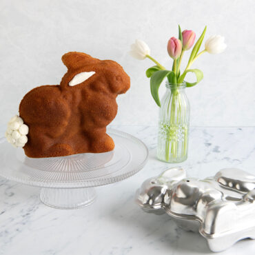 Nordic Ware aluminum 3D bunny cake pan with baked golden cake on stand beside spring tulips