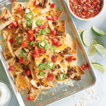 Two Nordic Ware aluminum half sheet pans with loaded nachos featuring melted cheese, jalapeños, and toppings