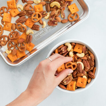 nordic ware aluminum jelly roll pan with snack mix, hand grabbing from a nearby bowl of snack mix