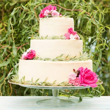 White frosted tiered wedding cake with garland and pink rose decorations, on cake stand, greenery in background