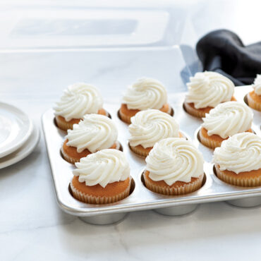 Nordic Ware aluminum 12-cavity muffin pan with frosted vanilla cupcakes, high domed lid in background
