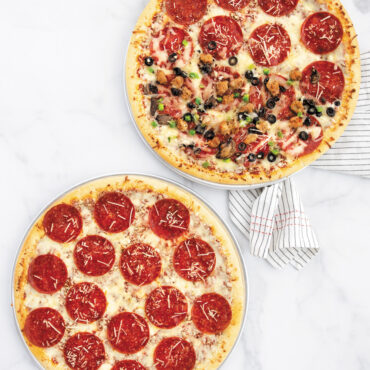 Two Nordic Ware aluminum pizza pans with baked pizzas - one traditional smooth, one perforated crisper pan