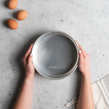 Hands holding Nordic Ware Naturals 8-inch round aluminum layer cake pan with natural silver finish and raised rim