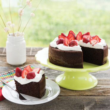 Chocolate layer cake with whipped cream and fresh strawberries on green cake stand, slice plated showing moist texture