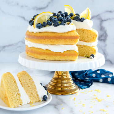 Two-layer lemon blueberry cake with white frosting, fresh blueberries and lemon slices, one slice cut and removed