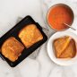Two cooked grilled cheeses on mini griddle, one grilled cheese plated with tomato soup