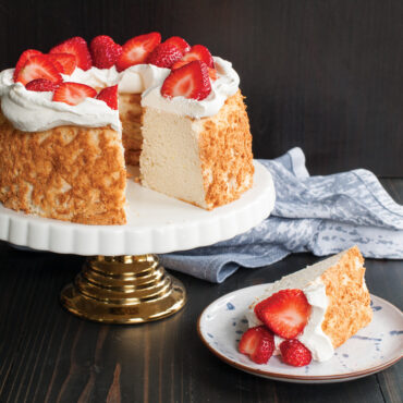 Golden angel food cake with whipped cream and fresh strawberry halves on white pedestal cake stand