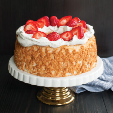 Golden angel food cake with whipped cream and fresh strawberry halves on white pedestal cake stand