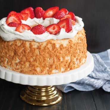 Baked angelfood cake with whipped topping and fresh strawberry halves on top, on cake stand