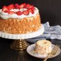 Large baked angelfood cake on cake stand with whipped cream and fresh strawberry garnish, mini cake with glaze and lemon zest on plate with fork
