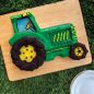 Baked tractor cake decorated in green and yellow frosting on wooden board on a fake grass surface. Plates in scene
