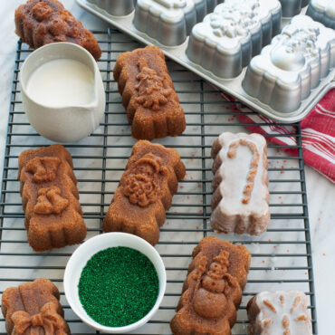 Six glazed holiday mini loaves with colorful sprinkles cooling on wire rack beside Nordic Ware aluminum mini loaf pan