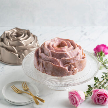 Rose Bundt Cake with pink frosting on clear cake stand with roses and pan placed around