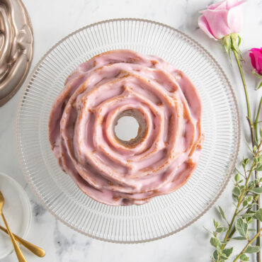 Rose Bundt Cake with pink frosting on clear cake stand with roses and pan placed around; overhead