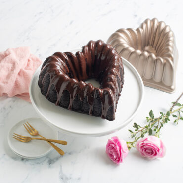 Elegant Heart Bundt cake with chocolate frosting on white cake stand, surrounded by pink roses and pan