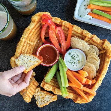 Pull apart cheese bread made in Stadium Bundt Pan, with dips and vegetables in the middle. Hand holding piece of bread and dipping.