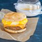 Microwaved egg with cheese and sausage patty on split biscuit set on parchment paper wrapper, product in background.