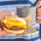 Hand holding microwaved egg with cheese and sausage patty on split biscuit set in parchment paper wrapper, on-the-go meal