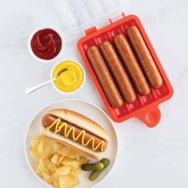 Nordic Ware aluminum hot dog steamer with cooked hot dogs, potato chips, and colorful condiment bowls arranged overhead