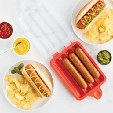 Nordic Ware hot dog steamer with cooked hot dogs in buns, served with chips and colorful condiments