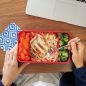 Bento Box filled with cooked carrots, pasta, chicken, and broccoli at a work desk with laptop