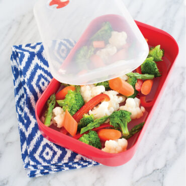 White Nordic Ware microwave steam cooker with cooked mixed vegetables, clear lid resting alongside