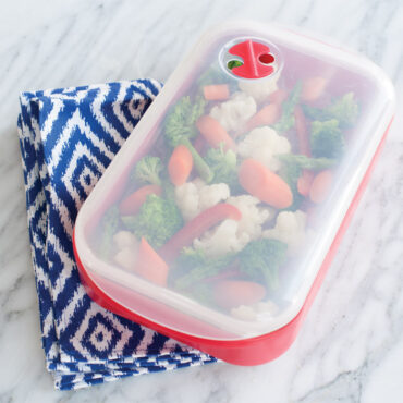 Nordic Ware aluminum steam cooker with perforated insert containing colorful steamed vegetables, clear lid partially visible